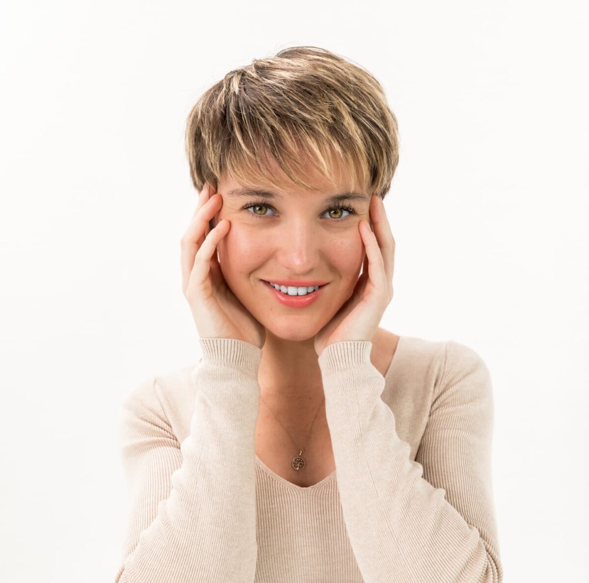 Woman with blonde pixie cut and hands on her cheeks smiles at camera.